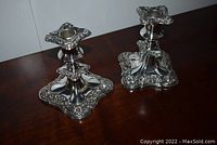 Pair of silver plated candle stick holders with ornate square bases and tops, showing floral and scroll decoration, sitting on wooden surface.