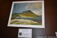 Photo showing the unframed print on table surface with visible full image depicting lake scene, signature, title, and artist name along with the printed artist profile card.