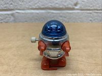 Front view of 1977 Tomy wind-up robot toy with blue dome head, silver body, orange arms and feet, and wind-up key on left side.
