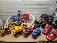 Multiple brightly colored plastic toy vehicles including construction dump trucks, off-road trucks, sports cars and a large Hot Wheels monster truck alongside a plastic playset with garage, ramps and a loop track.