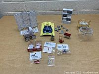 Photo showing assorted miniature dollhouse furniture and accessories on a gray surface. Items include white baby pram, decorative folding screen, grey cabinet, gold metal crib, white round table, red cushioned bench, miniature pots and pans, and multiple small packaged dollhouse miniature parts.