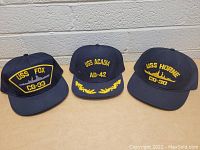Front view of three navy blue hats with yellow embroidered names USS Fox CG-33, USS Acadia AD-42, USS Horne CG-30.