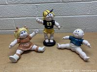 Front view of all three ceramic Cabbage Patch Kids figurines on wooden surface with white brick background showing girl, football player, and baby figurines.