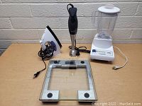 Photo of four assorted household and kitchen appliances on a wooden table, including a glass digital scale, white blender, black immersion blender, and white electric iron.