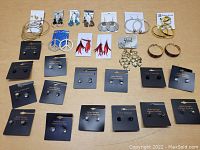 Overview of 27 pairs of earrings in various styles arranged sitting on a flat surface with some on display cards, including hoops, studs, dangles with feathers and peace signs.