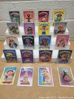 Front view of 20 vintage Garbage Pail Kids cards displayed in individual clear protective sleeves arranged in rows on white display stands against a white wall.