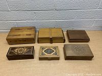 Lot of six vintage wooden boxes arranged in two rows on a beige surface against a brick wall, showing different wood finishes and decorative designs.