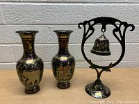 Front-facing photo of two metal vases and a decorative metal bell with a stand placed on a wooden surface against a white brick wall.