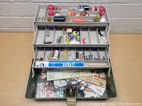 Photo of open vintage tackle box showing three-tiered trays with various fishing bobbers, lures, hooks, and weights.