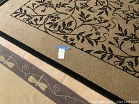 Photo showing details of large beige rug with black leaf vine design and part of smaller dragonfly rug beneath.