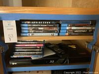 Photo of LG Blu-ray player with Blu-ray discs stacked above it inside a blue shelf. Titles visible include Upside Down, Death Race, Triage, Jordan Hex, and others.
