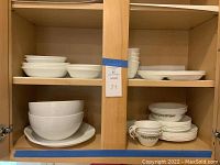 Upper and lower kitchen cabinet shelving filled with various Corelle dishes, including plain white bowls, plates, and cups with floral patterns.