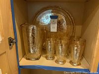 Pressed glass serving set displayed inside cabinet including tray, pitcher, and glasses with visible etched floral pattern.