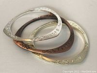 Three triple-sided metal bracelets in gold, copper, and silver tones, each engraved with 'Faith', 'Hope', or 'Love' and with delicate scrollwork designs on the edges, arranged stacked on a white surface.