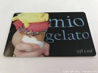 Close-up photo of the Mio Gelato gift card showing a hand holding a colorful gelato cone and the Mio Gelato logo on a black background.
