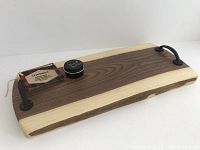 Wooden charcuterie board with natural and darker wood strips, black iron handles on each end, with attached D & DW Woodcraft tags and a small jar labeled 'Board Butter' on top.