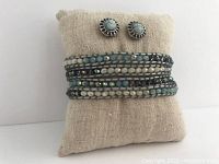 Front view of the leather wrap bracelet with mint, aqua, and pearl beads displayed on a beige cushion together with round aqua and silver stud earrings.