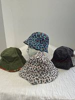 Four hats displayed on white surface with a glass head form, showing three reversible bucket hats with leopard and floral prints plus one black paisley Crooks Elite hat.