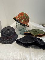 Four reversible summer bucket hats displayed on and around a glass head model: floral, olive green, black, and abstract patterns.