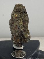 Front view of the raw chalcopyrite and pentlandite mineral specimen positioned upright on a black base with a Canadian quarter coin at the base for size reference.