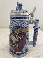Front view of ceramic stein depicting King Arthur in armor, details of castle turret lid and dagger handle with decorative patterns.