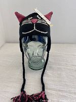 Front view of a black wool hat shaped as a smiling cat with pink ears and braided tassels displayed on a glass head form.