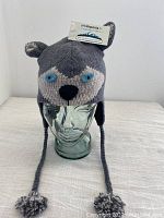 Front view of the grey animal knit wool hat displayed on a glass head model, showing knitted animal face with ears, braided tassels, and pom-poms.