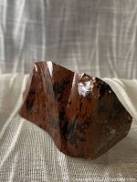 Chunk of mahogany obsidian volcanic glass on fabric background, showing reddish-brown and black colors from one angle.