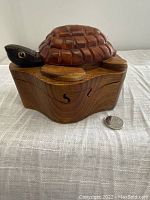 Side view of one wooden carved turtle puzzle stash box resting on fabric surface, showing carved segmented turtle shell lid and head with eyes, next to coin for scale.