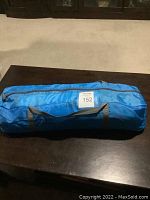 Blue fabric dome tent packed in a cylindrical carry bag with handles and zipper on a dark wooden table.