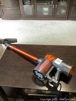 Full view of the vacuum cleaner laid on a dark surface showing the stick and handheld design with orange wand and grey components.