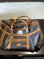 Front and side view of large brown leather travel duffle bag with tan leather trim and handles sitting on a dark surface.