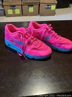 Pair of bright pink Nike Air Max 90 sneakers with blue midsole and black outsole display from an angle showing left shoe frontal and side view on dark wooden table.