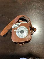 Fujifilm Instax Mini 25 instant camera with brown leather case and strap on dark surface, showing front with lens and flash.