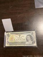 Front view of sealed Canadian 1 dollar banknote in heavy plastic protective case, showing Queen Elizabeth II and denomination.