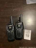 Image showing two black Uniden walkie talkies with antennas, each with an LCD screen and multiple buttons on the front, placed on a wooden surface alongside a MaxSold auction lot card