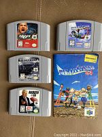 Four Nintendo 64 game cartridges (WCW Mayhem, Pilotwings 64, Forsaken 64, Madden 99) with Pilotwings 64 manual placed on a brown surface.