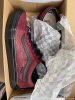 Top angle photo showing one shoe in an open box with checkered tissue paper, highlighting dark red suede and textured paneling with black sole and stripe.