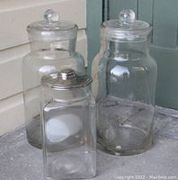 Photo of three large antique and vintage glass jars used in grocery or candy stores, showing clear glass containers with matching lids, two with glass lids and one with metal lid.