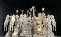 Photo showing eight large silver metal angel candlesticks arranged in a cluster, highlighting the detailed angel design and silver finish.