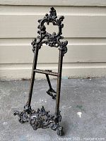 Front view of ornate brass folding picture easel frame standing upright on concrete surface with decorative scrollwork