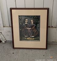 Framed antique hand coloured lithograph print "Two Girls" by Currier and Ives, 1863, shown standing against a wall with a wooden floor beneath.