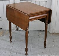 Front side view of wooden drop leaf table with one leaf lowered, dovetailed drawer and turned legs.
