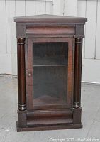 Full front view of the antique Victorian corner cabinet made of dark wood with glass door and decorative cylindrical columns on each side.
