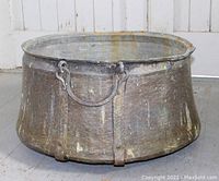 Front view of an enormous antique copper maple syrup cauldron showing weighty copper body, patina, incised designs, and metal handles attached on the sides.