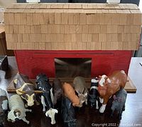 Photo showing handmade wooden toy barn with shingled roof and red painted walls. In front of the barn there is a collection of 11 vintage plastic farm animal figurines including bulls, cows, pig, donkey, sheep, and goat.