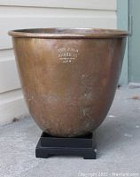 Copper stock pot marked 'The J. Van Range Co., Manufacturers, Ohio' with darkened patina and scratches, sitting on a small black stand.