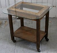 Full view of small antique oak rolling table with thick glass top and castered legs on wooden floor.