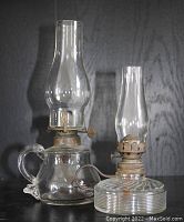 Two antique ship's finger oil lamps made of glass and metal, shown side by side against a dark background. One lamp has a taller chimney and a rounded glass reservoir with a handle; the other has a shorter chimney and a ribbed cylindrical glass reservoir with a metal handle ring.