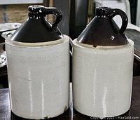 Photo of two large antique whiskey jug crocks showing dark brown tops with handles and light-colored bodies, placed on a wooden surface.